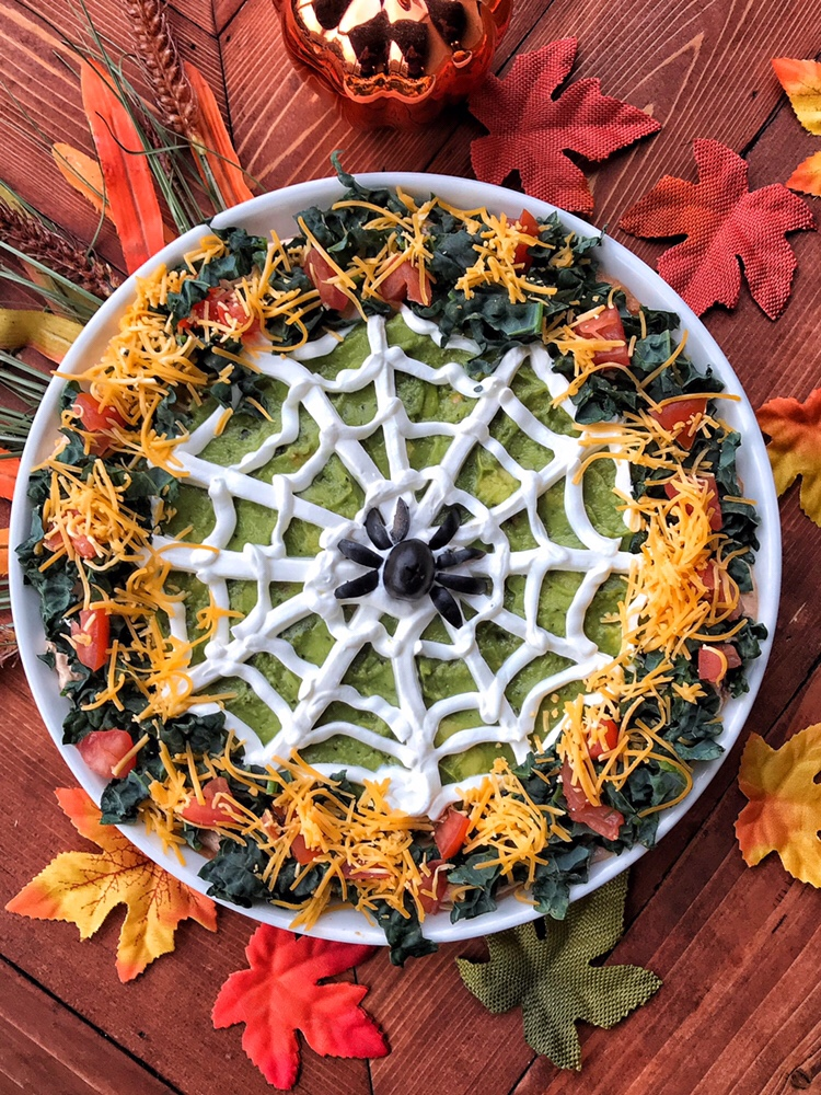 A Halloween guacamole dip with a sour cream spider web and an olive spider in the middle.