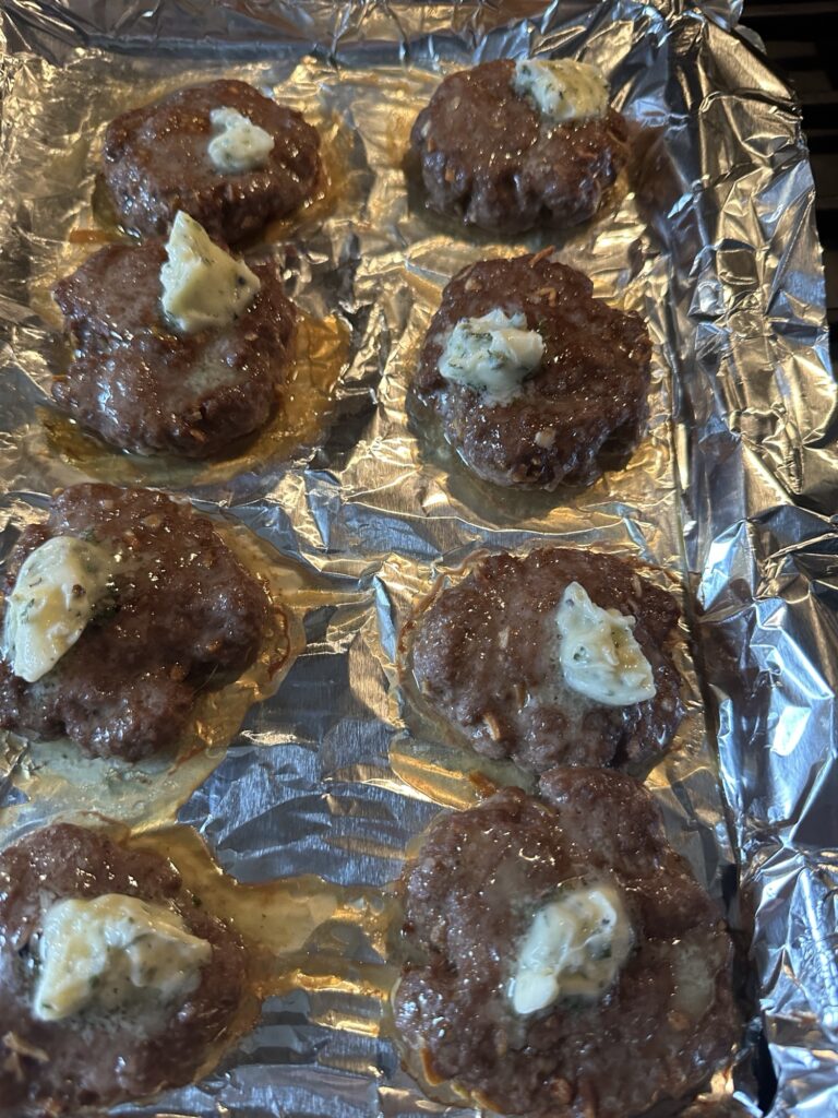 Cooked burger patties on a baking sheet with a dollop of butter mixture on each one.