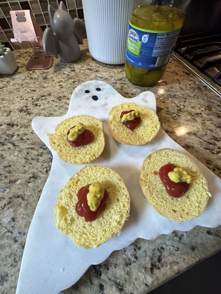 Four open faced buns with dollops of ketchup and mustard on a ghost shaped cutting board.