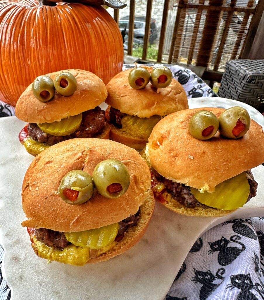 Four burgers with olive "eyes" in front of a pumpkin.