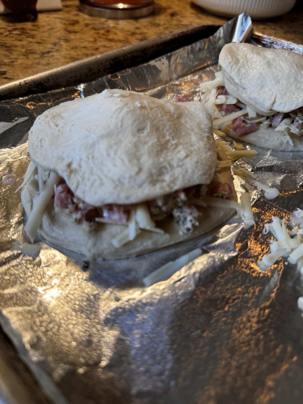 Two pieces of raw, round biscuit dough with ham and cheese in the middle of a baking tray.