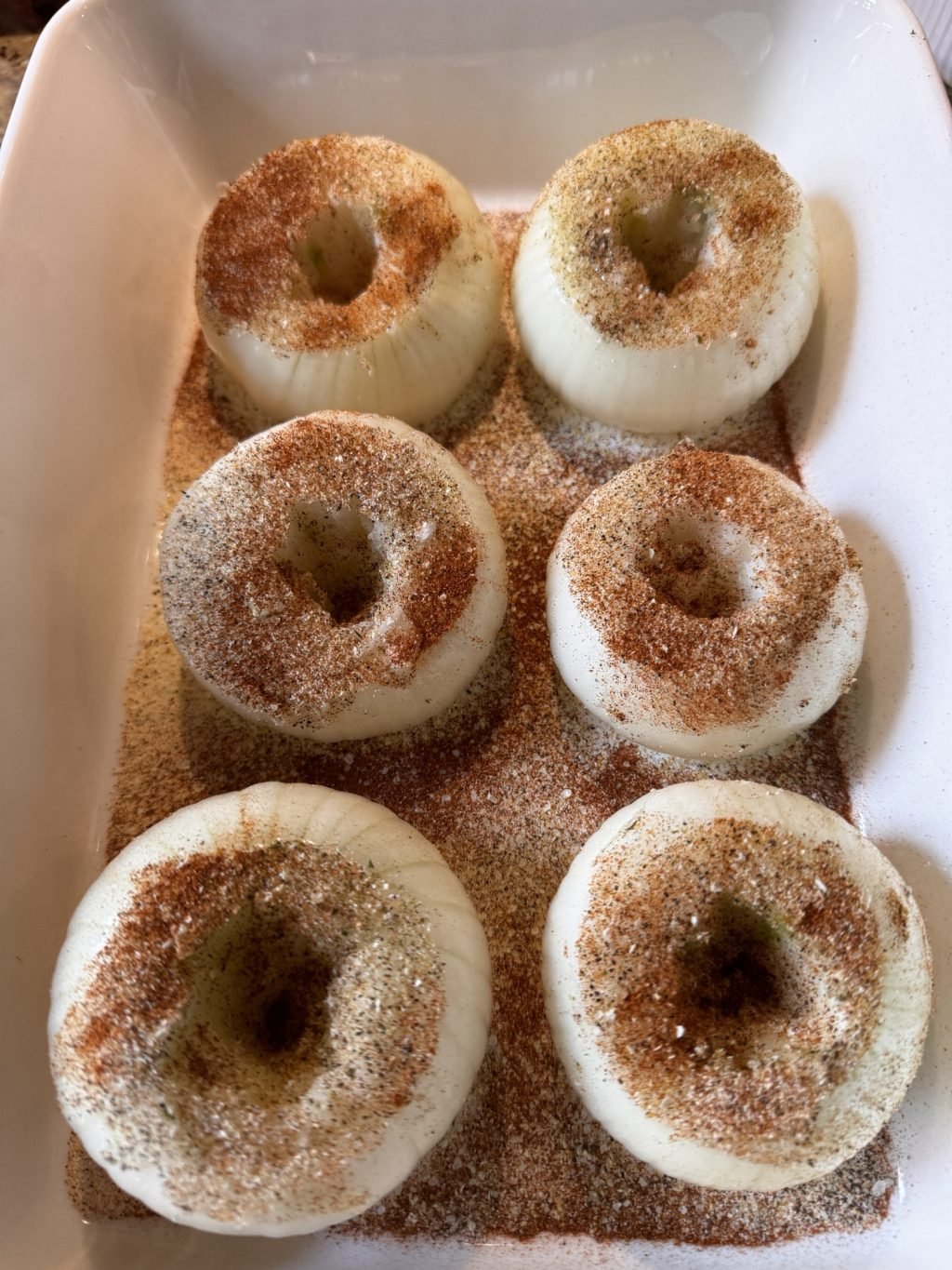 Six onions with holes hollowed out in the middle covered in spices in a baking dish.
