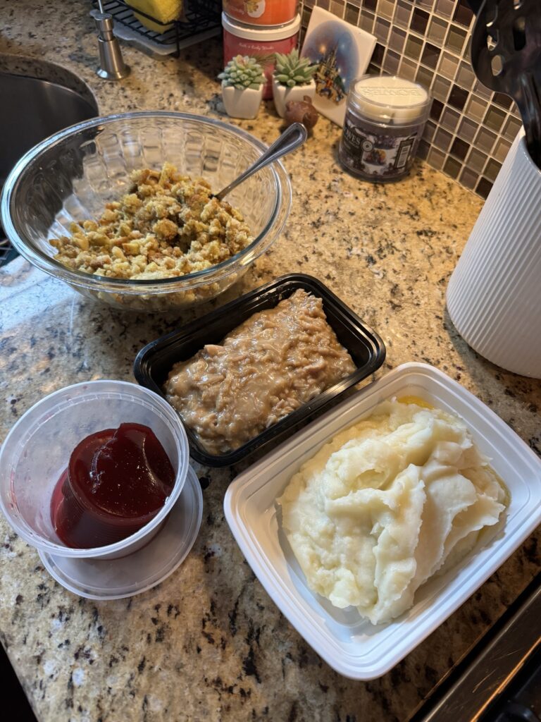 The ingredients for the Thanksgiving leftover sandwich on containers on the counter, including stuffing in a glass bowl with a metal spoon, a black plastic container of gravy, a white plastic container of mashed potatoes, and a clear plastic container of cranberry sauce.