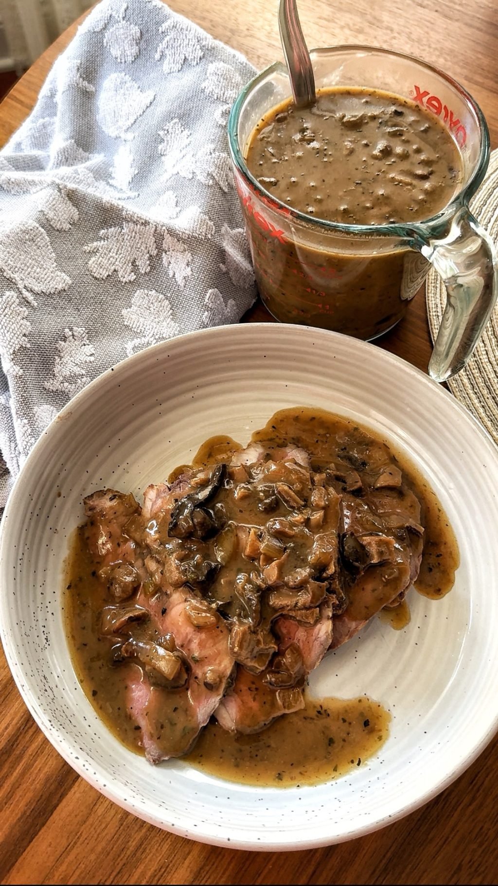A white plate of mushroom gravy on roast beef slices next to a glass measuring cup with more gravy and a spoon.