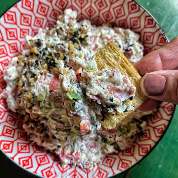 A Triscuit cracker with veggie dip on it is being held over a over a bowl of the dip.