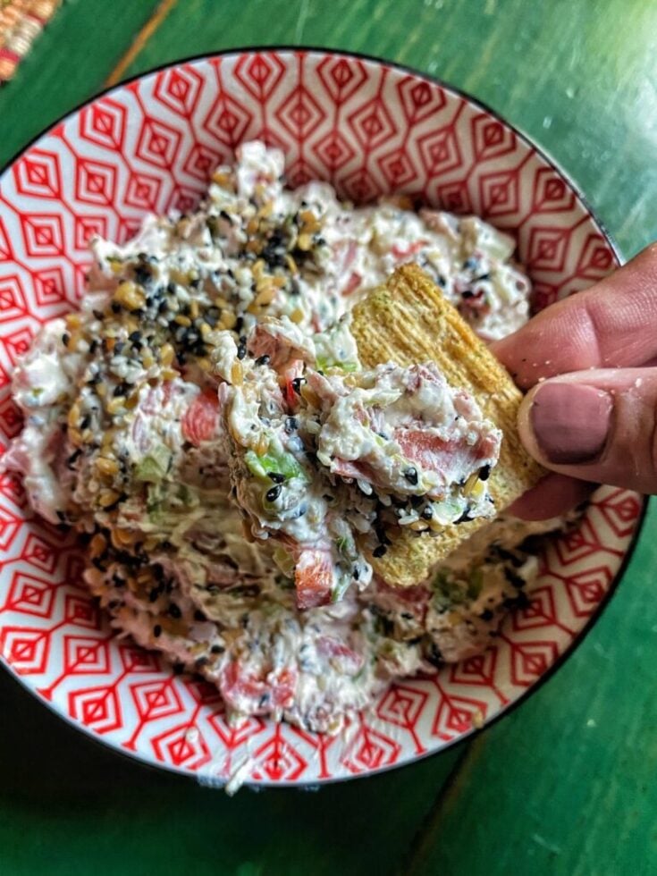 A Triscuit cracker with veggie dip on it is being held over a over a bowl of the dip.