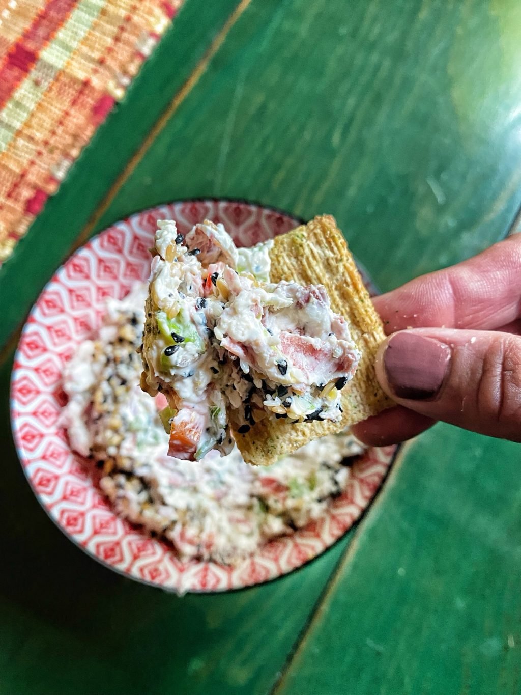 A Triscuit cracker with veggie dip on it is being held over a over a bowl of the dip.