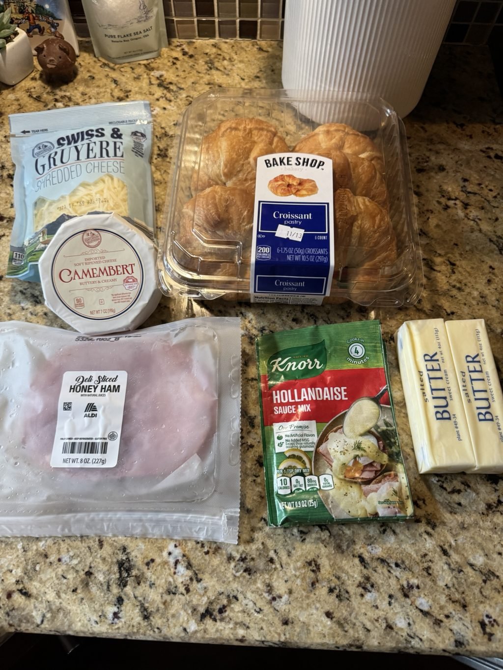 The ingredients for the baked croissant sandwiches on the kitchen counter including shredded cheese, camembert, croissants, deli ham, hollandaise mix, and two sticks of butter.