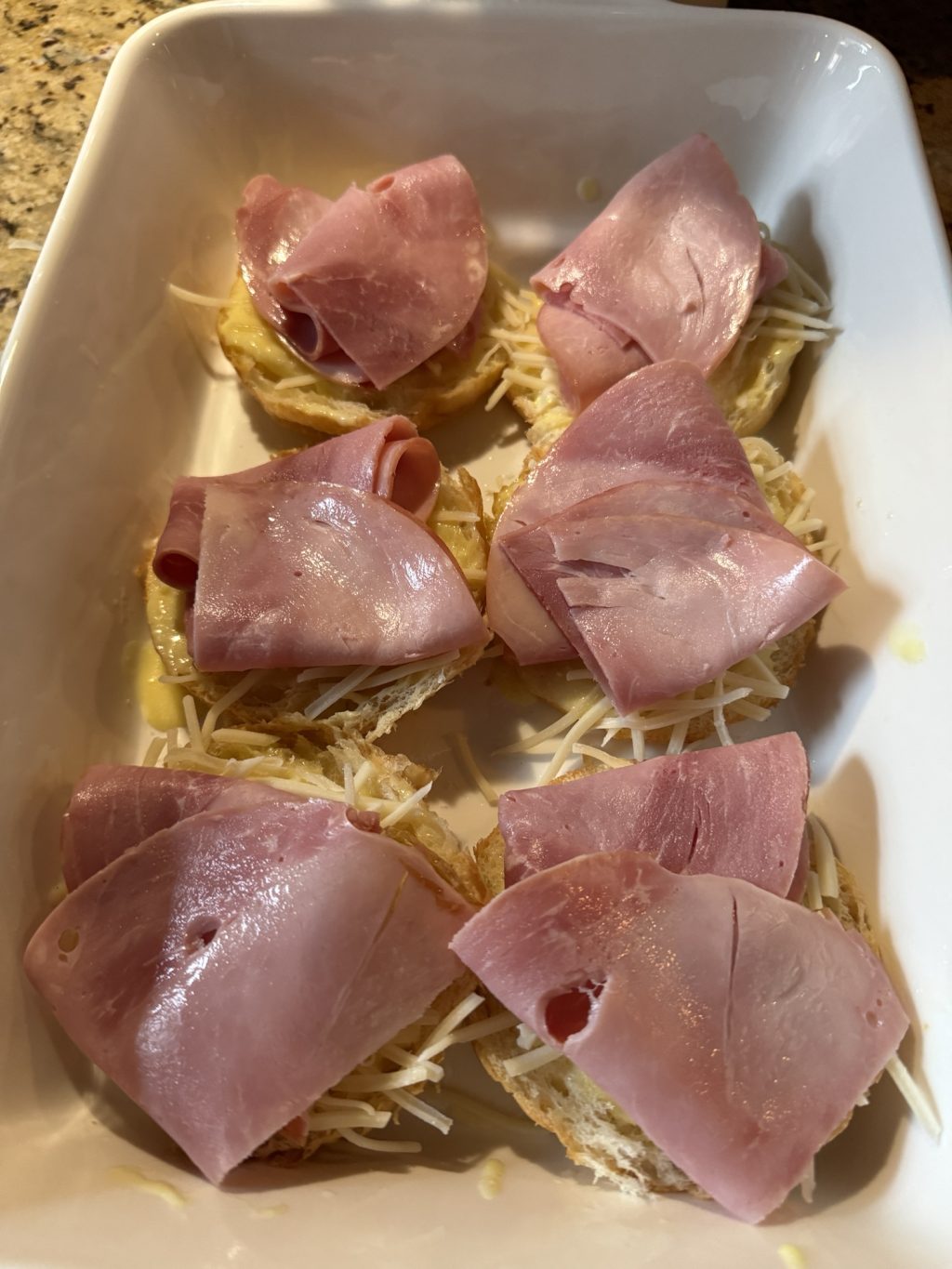 Sliced croissant halves in a baking dish topped with hollandaise and shredded cheese and sliced deli ham.