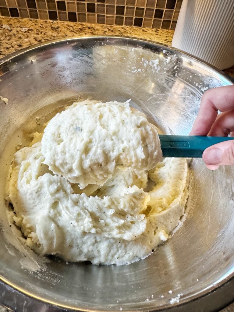 Mashed potatoes on a blue spatula held above a metal bowl filled with the rest of the mashed potatoes.