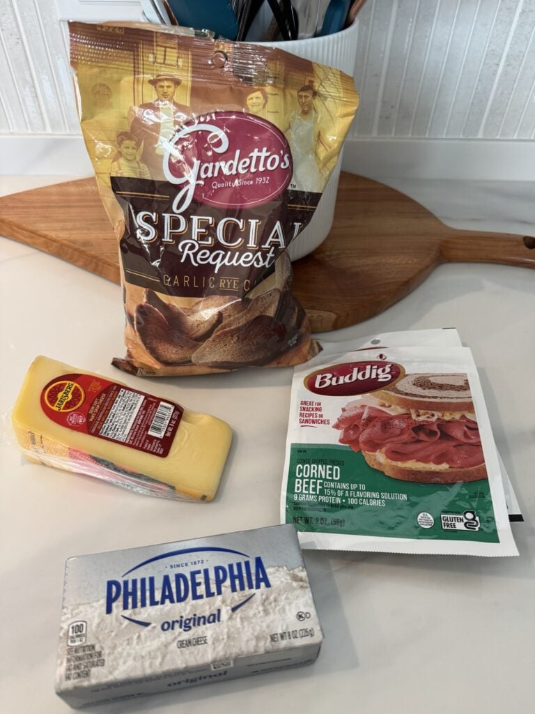 A bag of Gardetto rye chips, a wedge of Jarlsberg cheese, and a package of Buddig corned beef