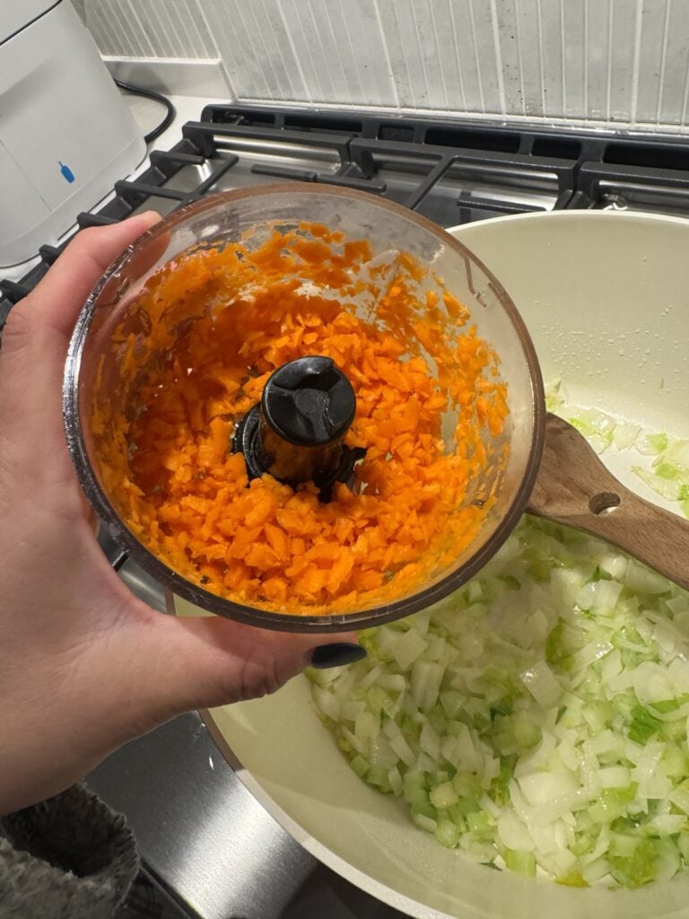 Food processor with carrots over diced greens and onion in the pan