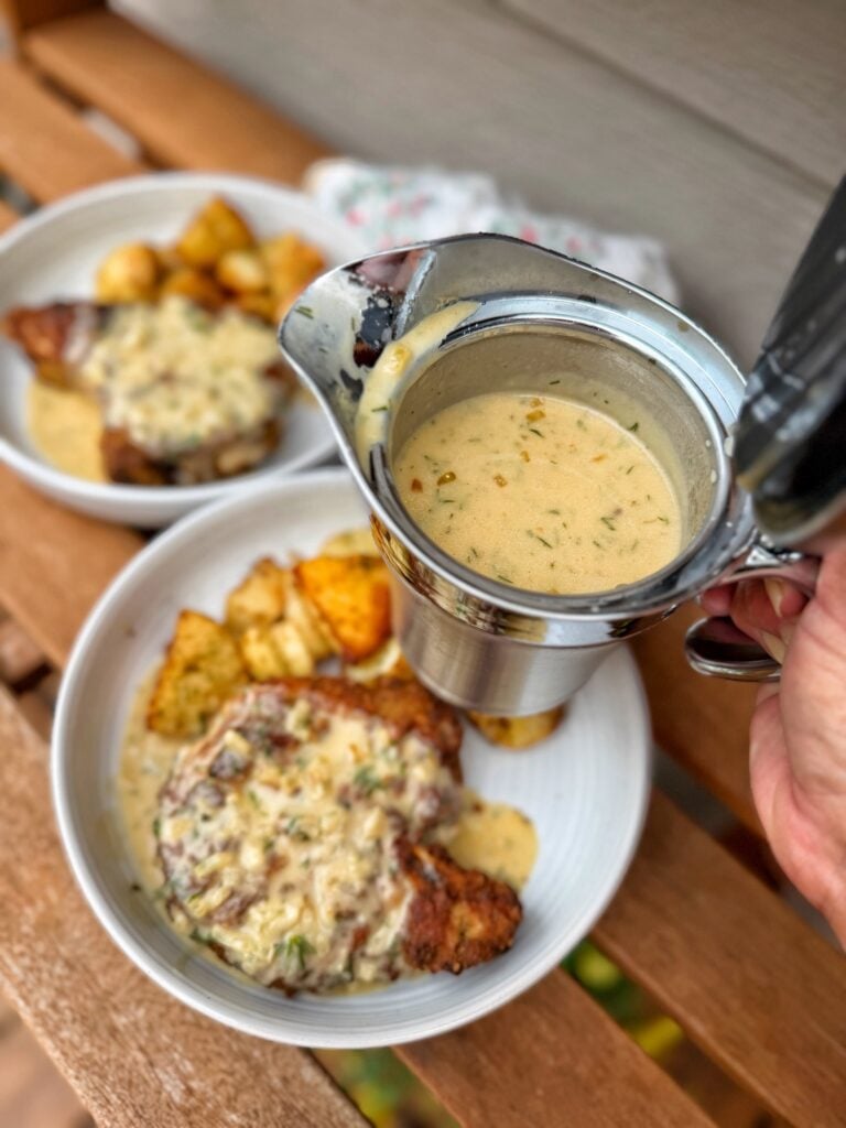 A metal gravy serving cup held over the finished pork chops and potatoes