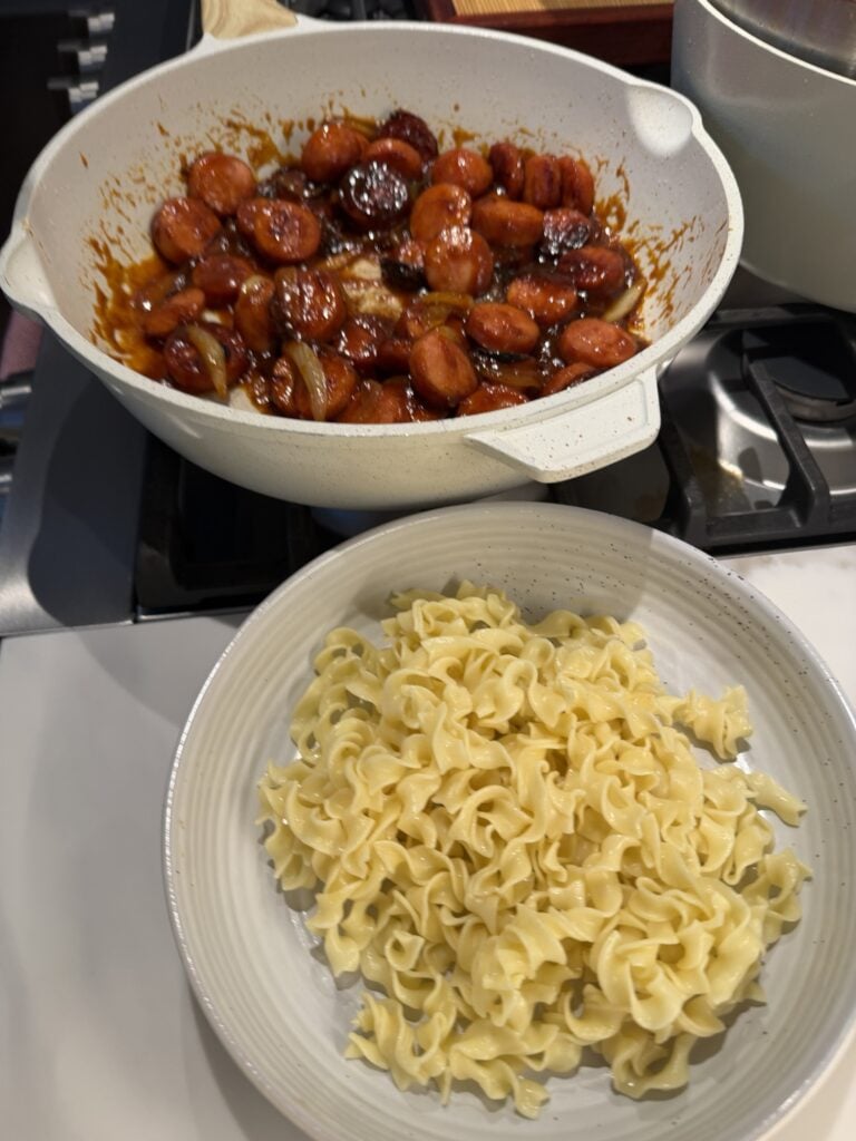 A plate of buttered noodles in front of a pot of saucy sausages