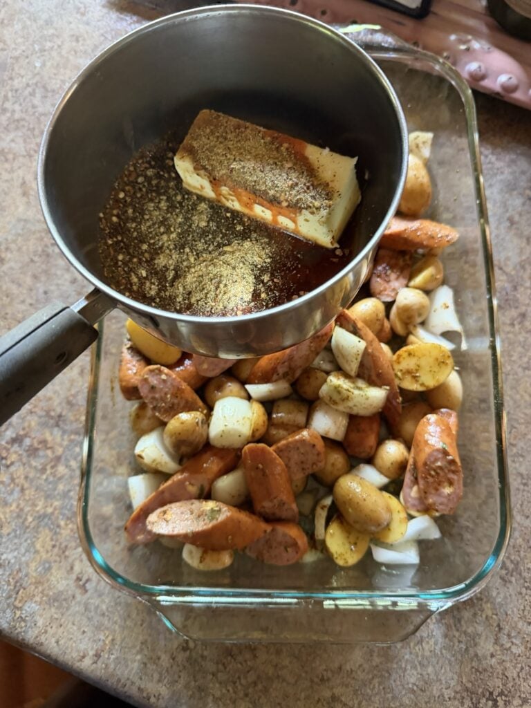 A pot with the ingredients for the sauce held over the baking dish of sausage, potatoes, and onions