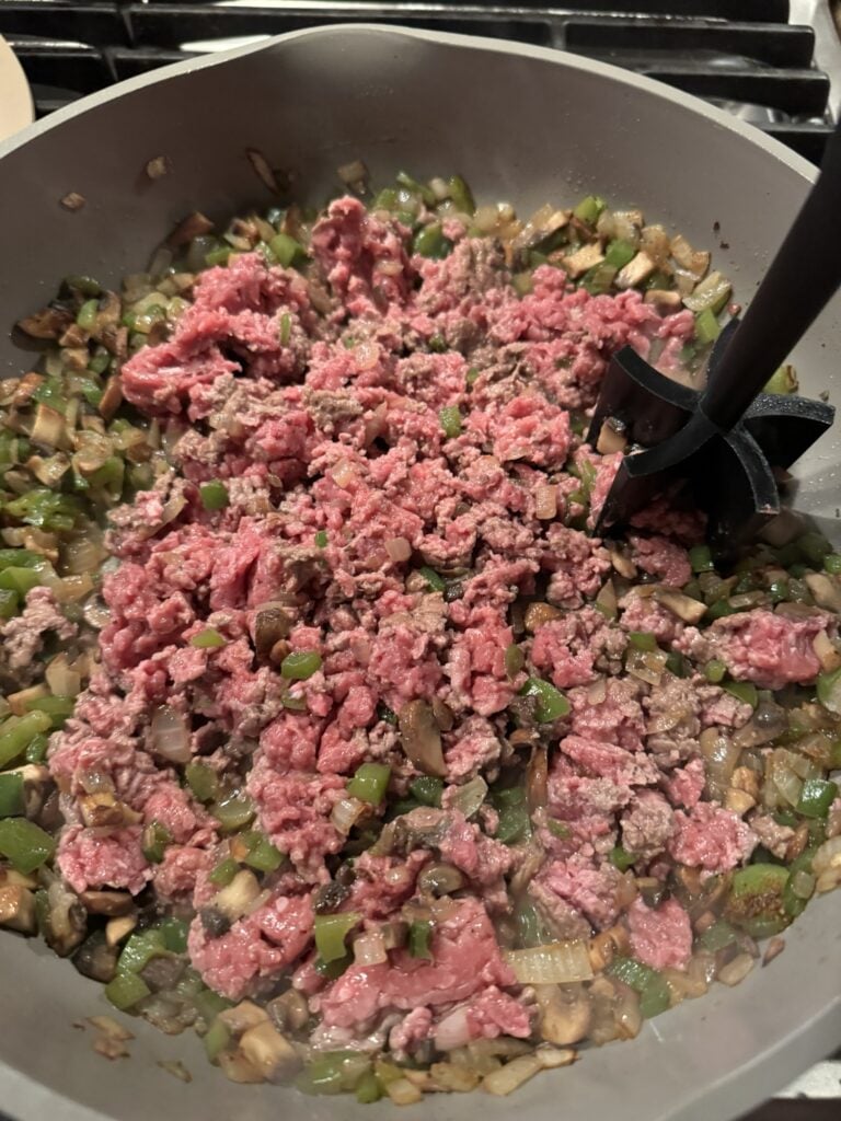 Ground beef being broken up by a meat chopper as it cooks with peppers onions and mushrooms