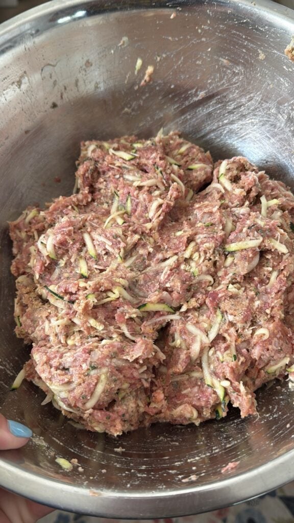 The meat, zucchini, and seasoning being mixed together in a metal bowl