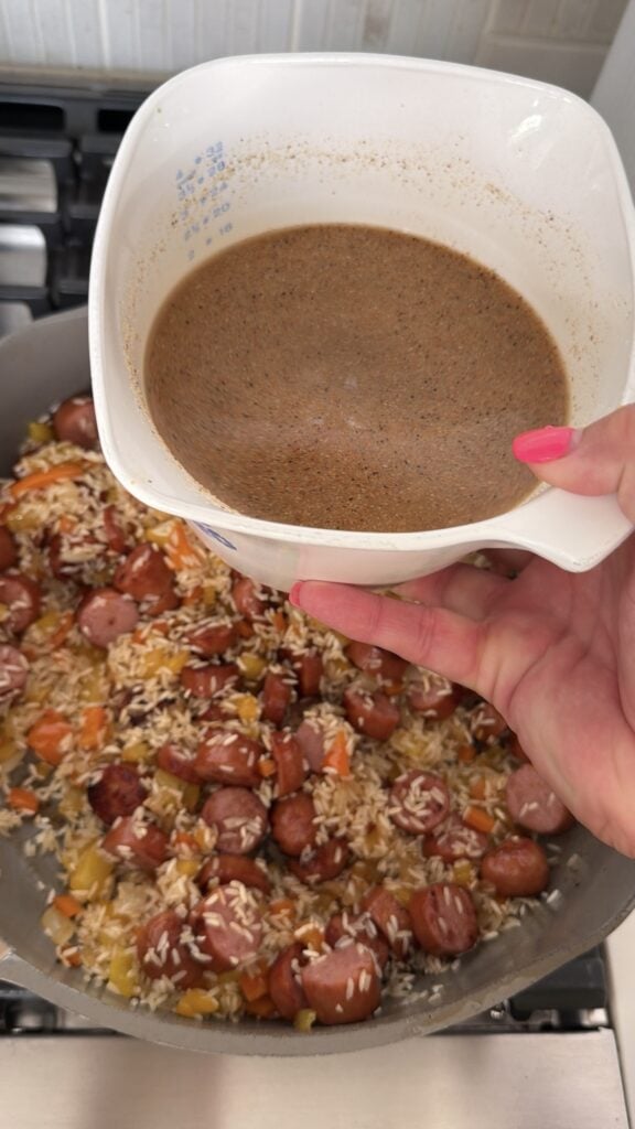 A container of broth about to be poured over the pan of sliced brats, rice, and pepper