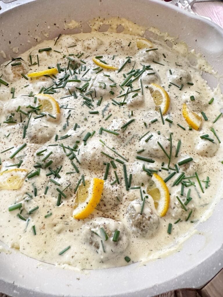 a pan of creamy white lemon meatballs topped with lemon slices and green chives sliced