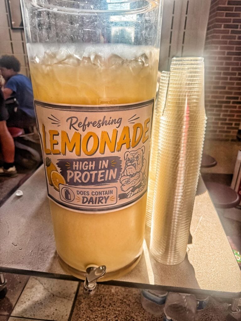 A lemonade urn at a party with a label that reads "Refreshing Lemonade, High in Protein, Does Contain Dairy"