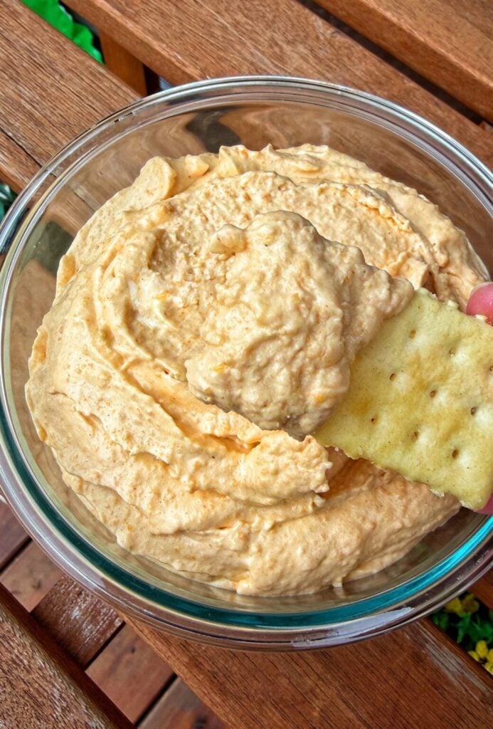 Creamy orange cheese spread in a glass bowl with a cracker dipped into it