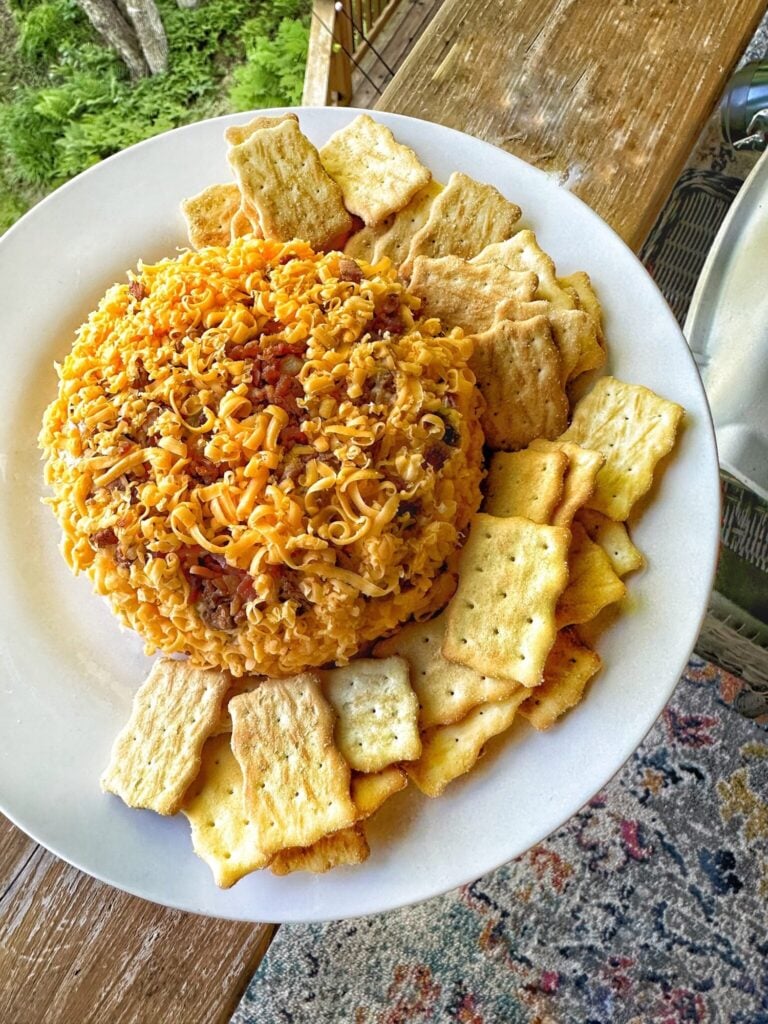 a cheese ball topped with cheddar cheese shreds with crackers on a white plate
