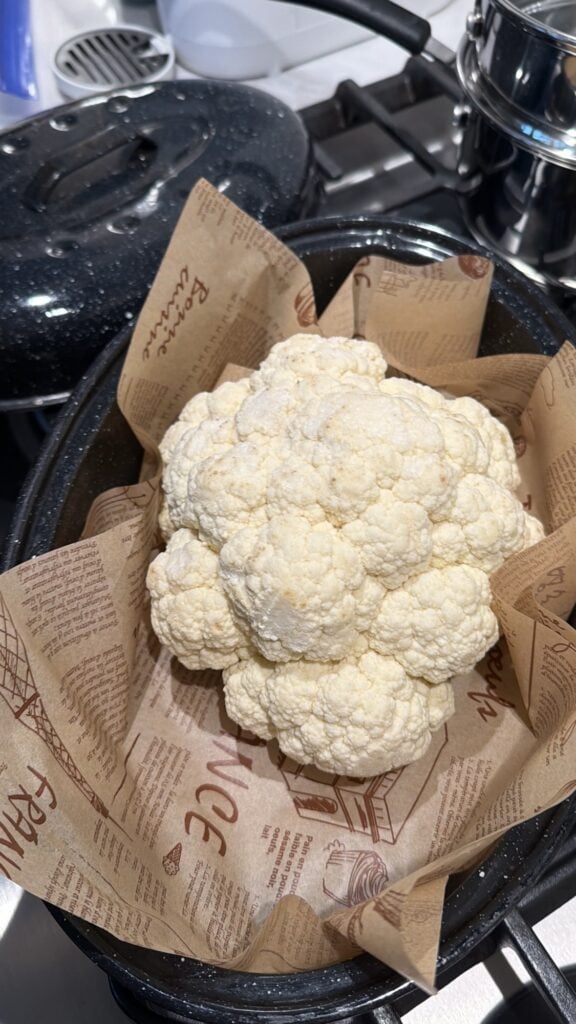 Whole cauliflower prepared for roasting in a covered black speckled roasting pan.