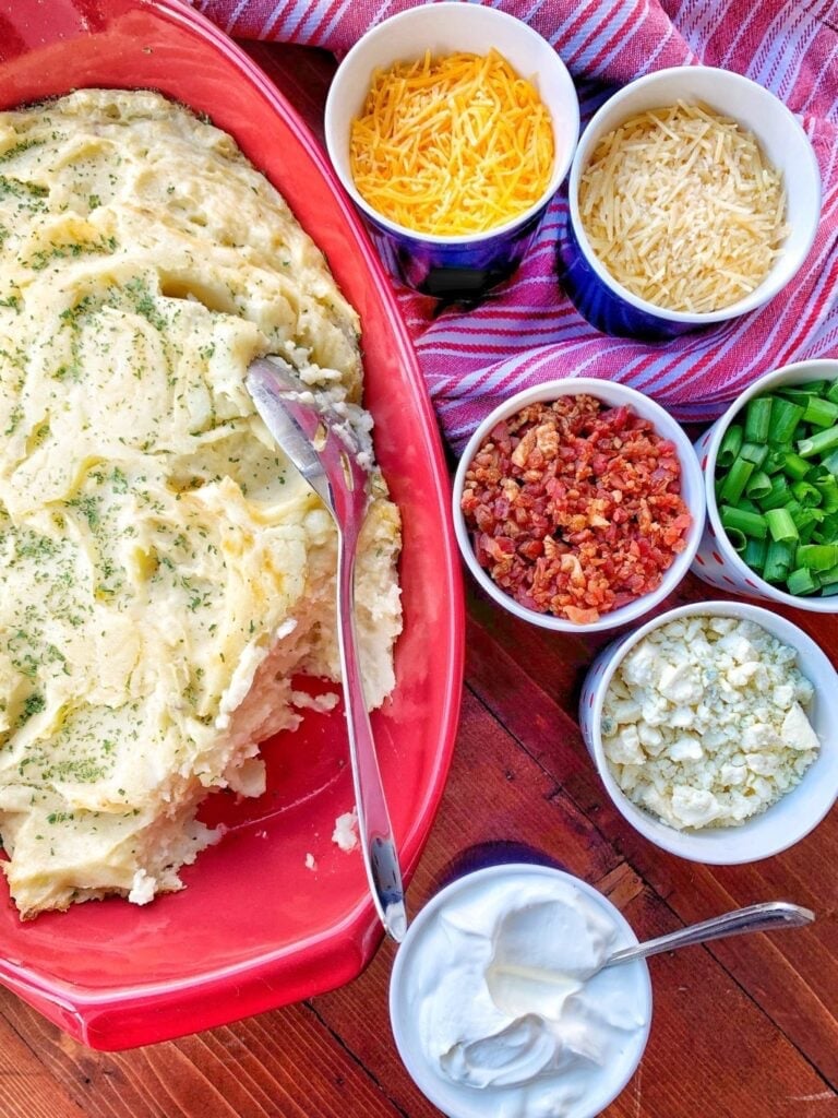 mashed potatoes in a casserole dish with smaller dishes filled with bacon, cheese, onions and sour cream