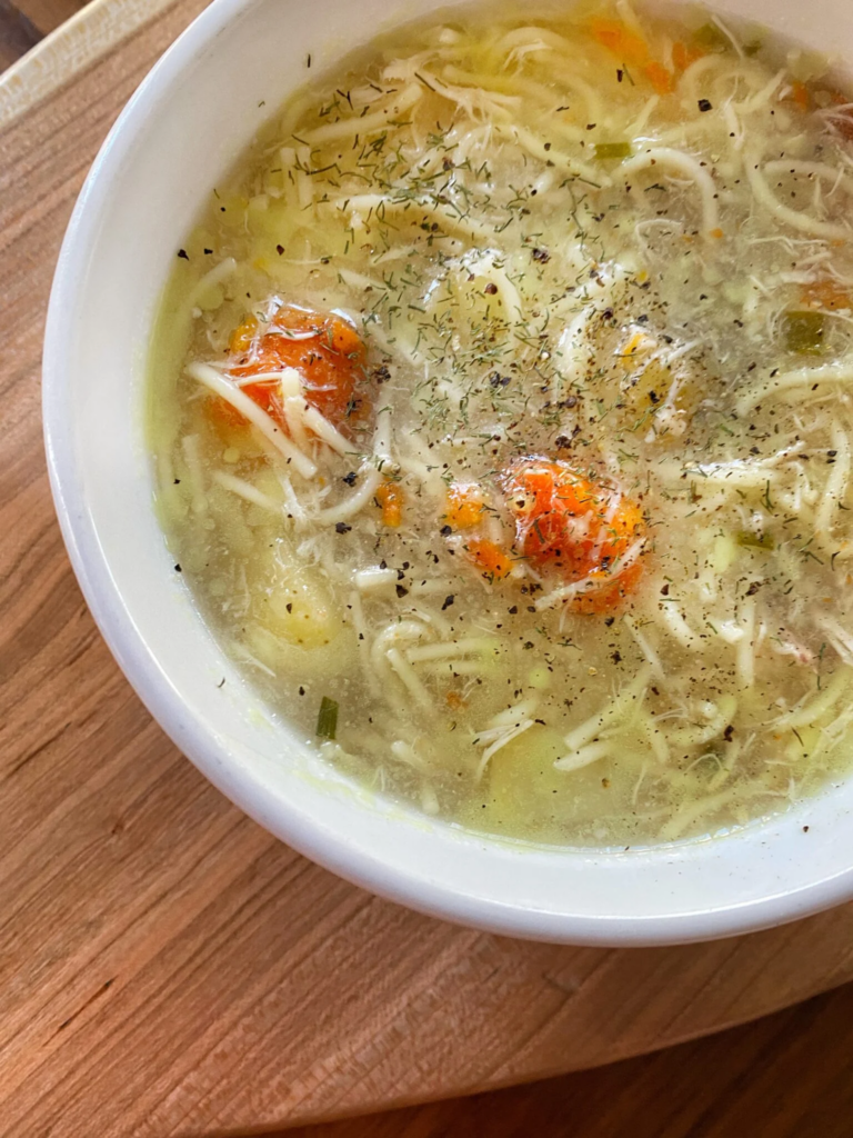 a bowl of chicken noodle soup with thin noodles, carrot pieces, dill and black pepper on top