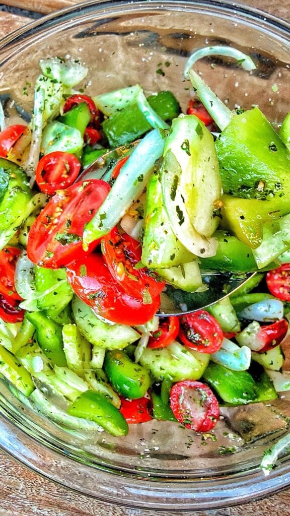 Close-up of garbage salad with halved tomatoes, green pepper chunks, cucumber, sweet onion, and dried herbs.