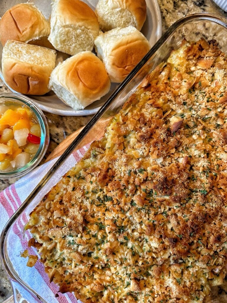 Baked chicken salad in a casserole dish with a crispy topping a side of rolls and fruit cocktail