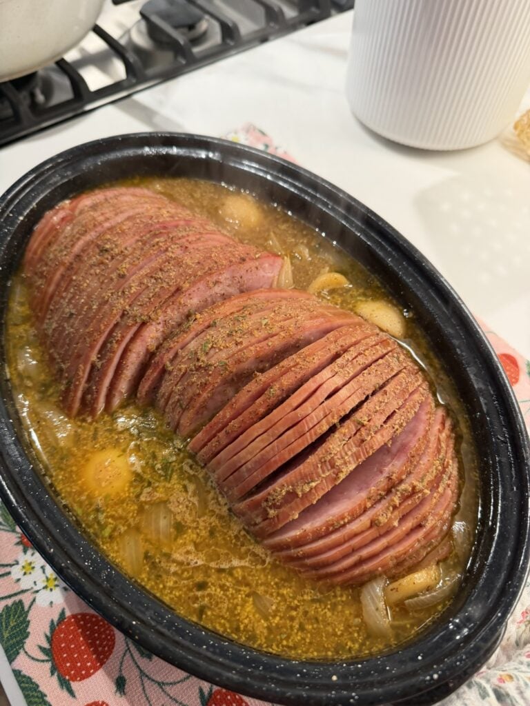 Sliced mini hams nestled among potatoes and onions with mustard sauce over the top.