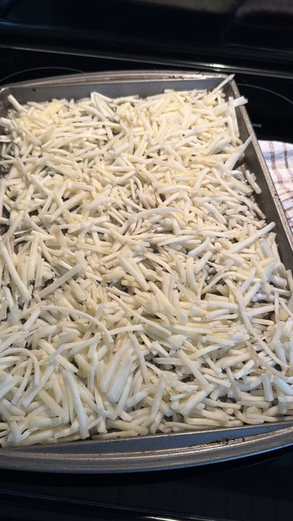 Thawed shredded hash browns spread in a thin layer across a buttered sheet pan.