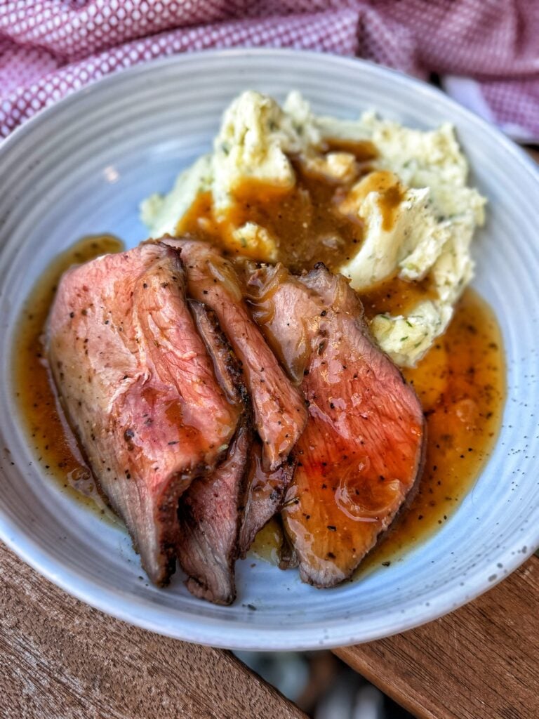 Thin slices of sirloin tip roast with onion gravy and chive mashed potatoes on a white plate.