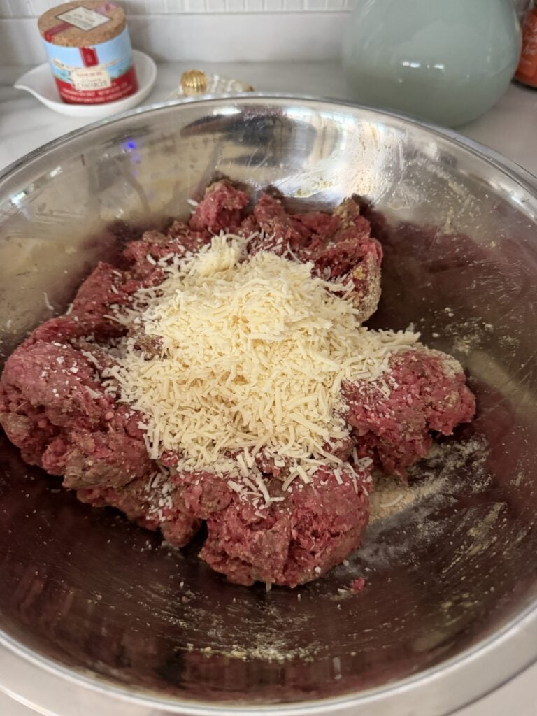 Ground beef topped with finely shredded Parmesan cheese and seasonings in a stainless steel mixing bowl.