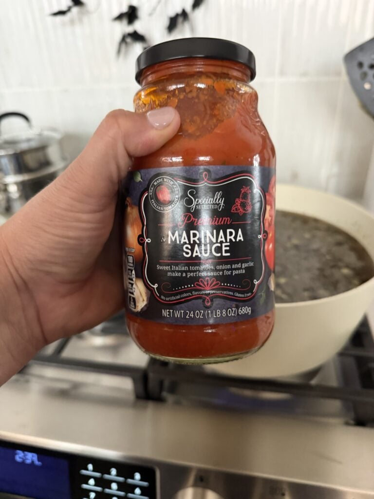 A jar of Specially Selected Premium Marinara Sauce held in front of a pot on the stove.