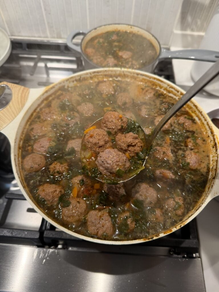 A ladle of mini meatballs, spinach, and diced vegetables held over a pot of mini meatball soup.